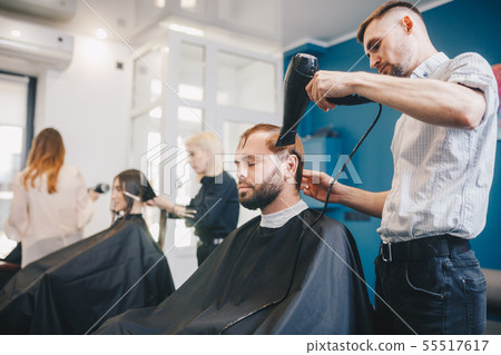 young bearded man getting groomed hairdresser with hair dryer barbershop. Professional Hairdresser young bearded man getting groomed hairdresser with hair dryer barbershop. Professional Hairdresser 55517617