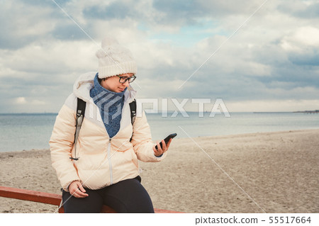 Caucasian woman in hat and jacket with a backpack in winter sits on a wooden pier on the beach near 55517664