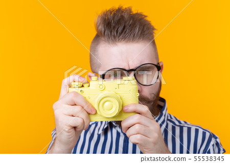 Cute young male photographer with a mustache and beard is photographing a yellow vintage camera on a 55538345