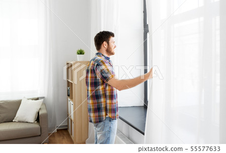 young man opening window curtain at home 55577633