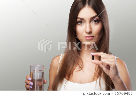 Pretty young woman who takes a black pill and holds a glass of water. Isolated on a gray background 55709495