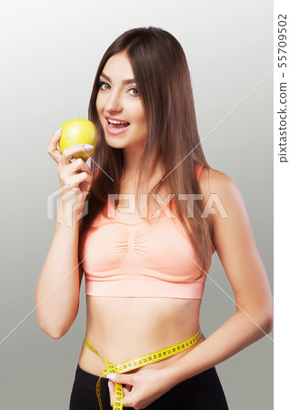 A young woman measures her waist with a tape measure and eats an apple. Strict diet. Beautiful body A young woman measures her waist with a tape measure and eats an apple. Strict diet. Beautiful body 55709502