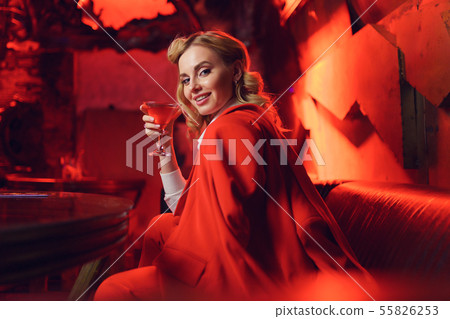 Photo of happy blonde in red jacket looking at camera with cocktail in her hand in nightclub 55826253