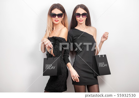 Black friday. Two women wearing black dress and sunglass, with shopping paper bag, look at camera in Black friday. Two women wearing black dress and sunglass, with shopping paper bag, look at camera in 55932530