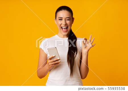 Gorgeous Woman Gesturing OK And Holding Cellphone On Yellow Background Gorgeous Woman Gesturing OK And Holding Cellphone On Yellow Background 55935538