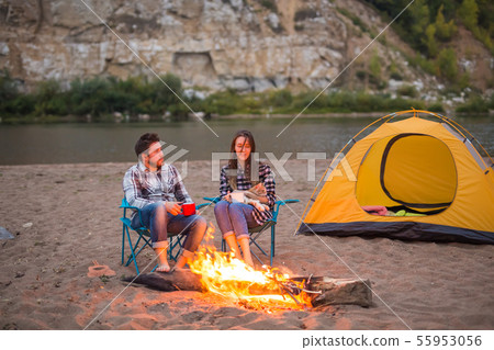 People, tourism and nature concept - Romantic evening, man embrace woman sitting near a campfire People, tourism and nature concept - Romantic evening, man embrace woman sitting near a campfire 55953056