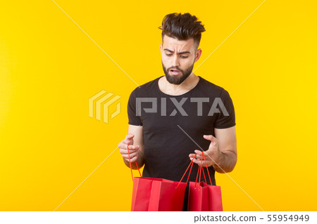 Cute young sad bearded man looks at the purchase in shopping bags on a yellow background with copy Cute young sad bearded man looks at the purchase in shopping bags on a yellow background with copy 55954949