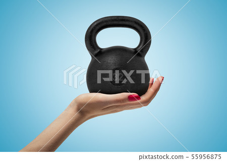 Side closeup of woman's hand facing up and holding big black 32 kg kettlebell on light blue gradient 55956875