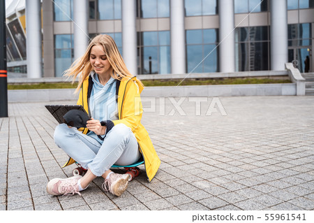 A beautiful blonde girl sits on a street and uses a tablet. 55961541