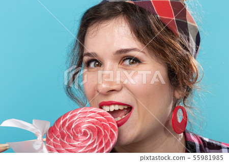 Close up portrait of beautiful young woman eating lollipop over blue background Close up portrait of beautiful young woman eating lollipop over blue background 55981985