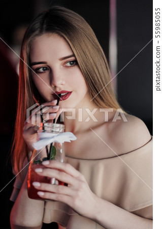 Young beautiful brown-haired girl in a nightclub with neon lights sits by the glass and drinks a red 55985055