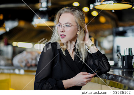 A young beautiful blond woman with glasses sits at the bar in a yellow interior. Drinks alcoholic A young beautiful blond woman with glasses sits at the bar in a yellow interior. Drinks alcoholic 55986162