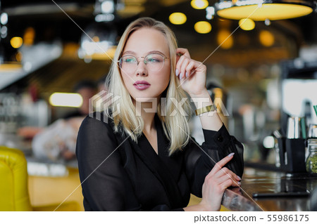 A young beautiful blond woman with glasses sits at the bar in a yellow interior. Drinks alcoholic A young beautiful blond woman with glasses sits at the bar in a yellow interior. Drinks alcoholic 55986175