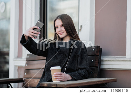 Young beautiful millenial girl in a coat is sitting on an outdor bench with a paper cup of coffee Young beautiful millenial girl in a coat is sitting on an outdor bench with a paper cup of coffee 55986546