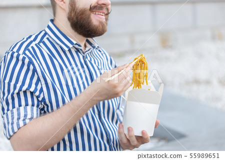An unidentified young male hipster with a mustache and beard is eating chinese noodles with wooden 55989651