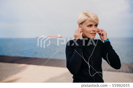 A portrait of young sportswoman with earphones standing outdoors on beach. 55992035