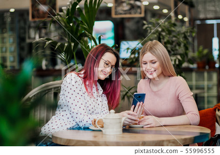 Two young beautiful girls sit in a green cafe at a table. Drink tea with croissants, chatting 55996555