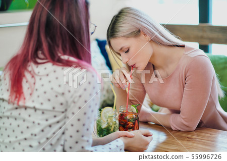 Two young beautiful girls, a blonde in a pink dress and a brunette with dyed hair, sit at a cafe Two young beautiful girls, a blonde in a pink dress and a brunette with dyed hair, sit at a cafe 55996726