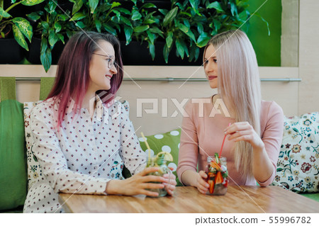 Two young beautiful girls, a blonde in a pink dress and a brunette with dyed hair, sit at a cafe 55996782
