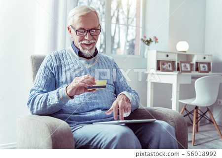 Grey-haired retired man holding bank card while shopping online Grey-haired retired man holding bank card while shopping online 56018992