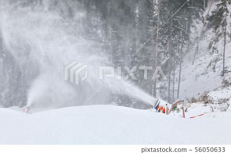 Orange and blue snow making cannon spreading ice crystals over ski piste, trees in background Orange and blue snow making cannon spreading ice crystals over ski piste, trees in background 56050633