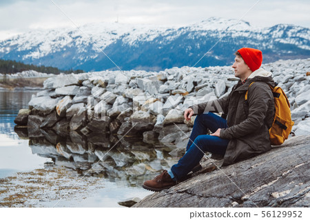 Traveler man with a yellow backpack wearing a red hat sitting on the shore on the background of 56129952