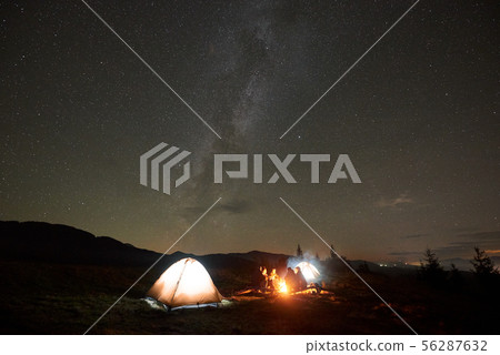 Group of tourists with guitar by burning bonfire under dark starry sky with Milky Way constellation. 56287632