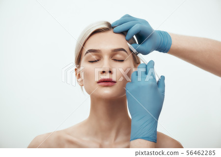 Cosmetic injection. Portrait of young pretty woman keeping eyes closed while doctors hand in blue Cosmetic injection. Portrait of young pretty woman keeping eyes closed while doctors hand in blue 56462285