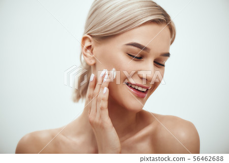 Portrait of cute blonde woman spreading cream on her face and smiling while standing against grey 56462688