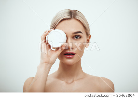 Deep hydration. Portrait of cute young blonde woman showing face cream while standing against grey Deep hydration. Portrait of cute young blonde woman showing face cream while standing against grey 56462804