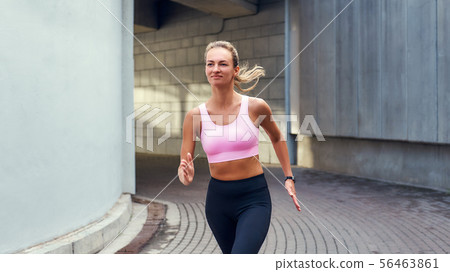 Enjoying run. Young smiling woman in comfortable sports clothing is running outdoors along the Enjoying run. Young smiling woman in comfortable sports clothing is running outdoors along the 56463861