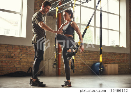 My personal trainer. Confident young man is showing slim athletic woman how to train legs with Trx 56466152