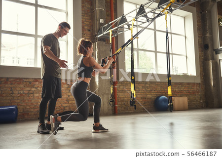 Postive workout. Beautiful young woman in sportswear doing trx exercises at gym while her personal Postive workout. Beautiful young woman in sportswear doing trx exercises at gym while her personal 56466187