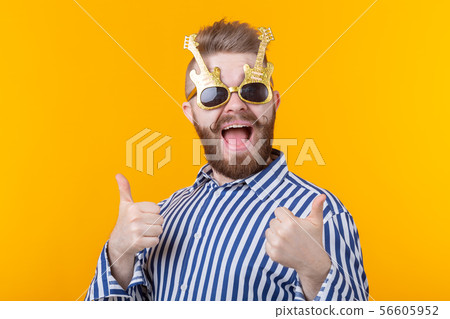 Joyful hipster young man raising his thumbs up in glasses with guitars posing against a yellow Joyful hipster young man raising his thumbs up in glasses with guitars posing against a yellow 56605952