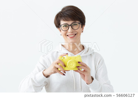 Positive middle-aged woman in glasses holds in hand a vintage yellow camera posing on a white 56606127