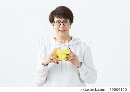 Positive middle-aged woman in glasses holds in hand a vintage yellow camera posing on a white 56606130