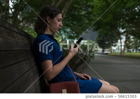 Side view of a young woman sitting on a bench in park holding her smartphone Side view of a young woman sitting on a bench in park holding her smartphone 56615303