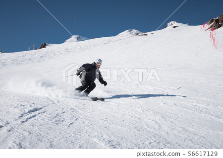 A bearded mature aged male skier in a black ski suit descends along the snowy slope of a ski resort A bearded mature aged male skier in a black ski suit descends along the snowy slope of a ski resort 56617129