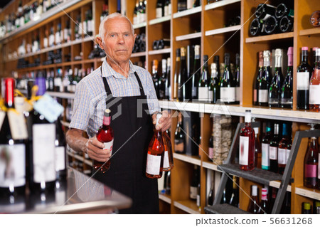 Confident elderly salesman of wine house arranging wine bottles on shelves rack Confident elderly salesman of wine house arranging wine bottles on shelves rack 56631268