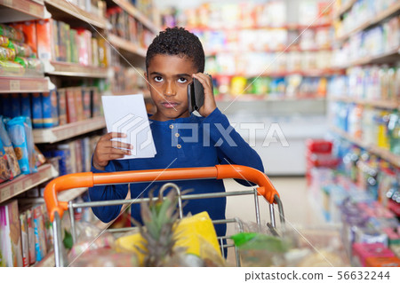 Focused African tween boy talking on phone and looking at shopping list while visiting supermarket 56632244