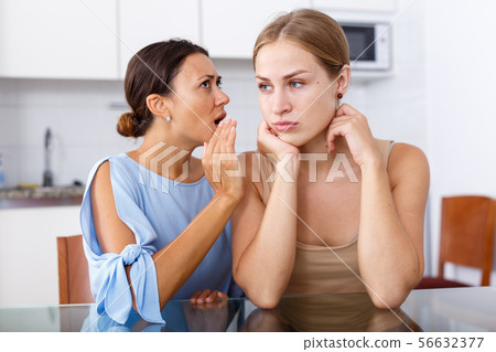 Upset young women friends whispers in the ear and talking at table in home Upset young women friends whispers in the ear and talking at table in home 56632377