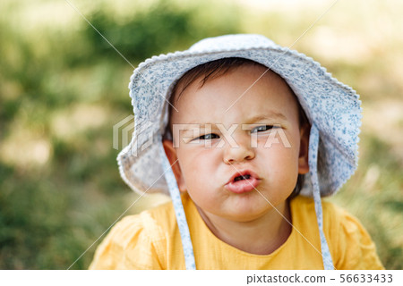 Caucasian baby girl is grimacing to the camera in hat and yellow dress. Summer childhood Caucasian baby girl is grimacing to the camera in hat and yellow dress. Summer childhood 56633433