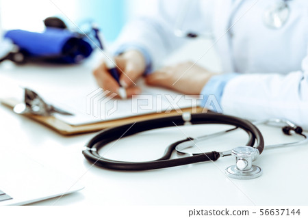 Closeup of stethoscope. Female doctor fills up medical form while sitting at the desk in hospital 56637144
