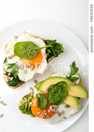 Fried eggs on a toasts bread with avocado, spinach and seeds on a white plate on the white 56642656