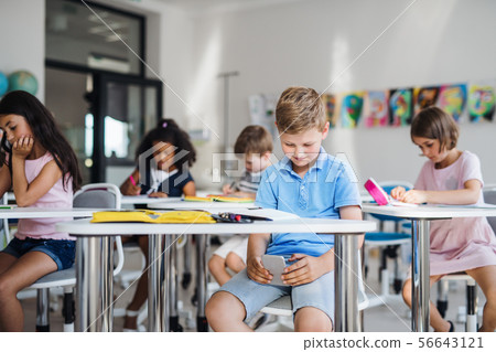A small school boy with smartphone sitting at the desk in classroom, playing. A small school boy with smartphone sitting at the desk in classroom, playing. 56643121