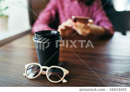 Young woman in the background scrolls smartphone, cup of coffee in front. Young woman in the background scrolls smartphone, cup of coffee in front. 56658405