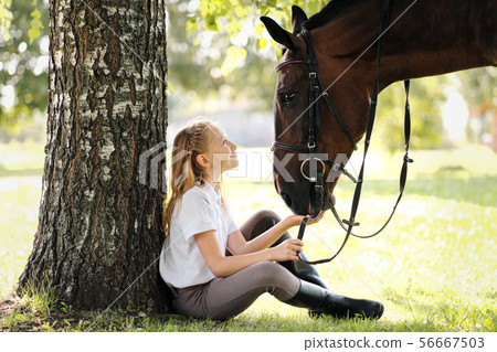 Girl teenager jockey sits in a green clearing under a tree. Feeds a horse an apple and strokes it. 56667503