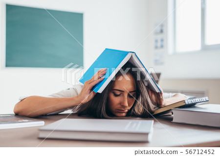 Tired female student sitting at the table with a book on her head. 56712452