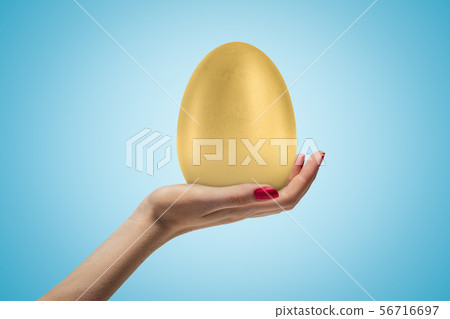 Female hand holding big golden egg on blue background Female hand holding big golden egg on blue background 56716697