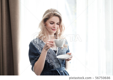 Beautiful young blonde woman in a blue robe by the window. Drinks coffee or tea from a white cup Beautiful young blonde woman in a blue robe by the window. Drinks coffee or tea from a white cup 56750686